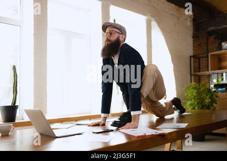 Young bearded man, office clerk having fun, doing yoga on wooden table in modern office at work time with gadgets. Concept of business, healthy Stock Photo