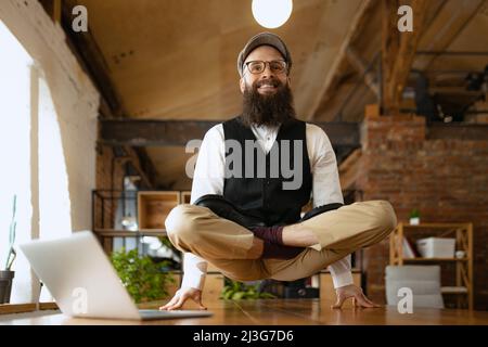 Emotional office clerk, young man doing yoga on wooden table in modern office at work time with gadgets. Concept of business, healthy lifestyle, sport Stock Photo