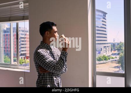 A young businessman drinks his coffee and looks out on the cityscape from office Stock Photo