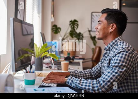 A young Asian businessman types at his desk on the computer while smiling Stock Photo