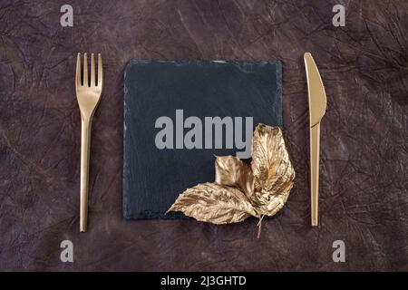 golden knife and fork on brown leather texture next to a black stone tile Stock Photo