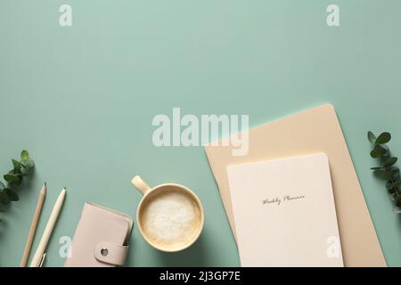 Aesthetic minimalist style home office desk table with notebooks, cup of coffee, feminine accessories and eucalyptus leaves on green background. Flat Stock Photo