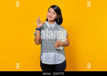 Beautiful Asian woman holding laptop smiling and showing okay sign over yellow background Stock Photo