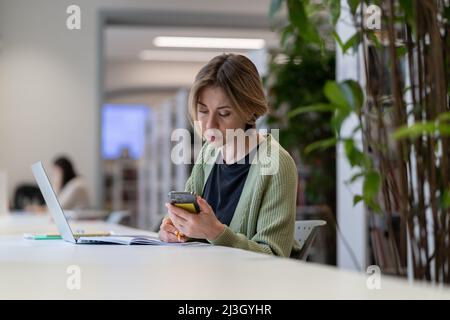 Focused female university teacher using laptop while sitting in quiet cozy library Stock Photo