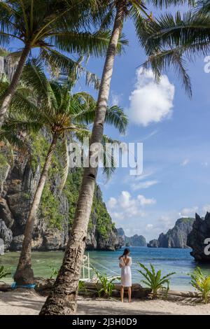 Philippines, Palawan, El Nido, Bacuit Archipelago, Miniloc Island, Secret Lagoon, young Filipino woman on white sand beach, under tall coconut trees, surrounded by karst cliffs Stock Photo