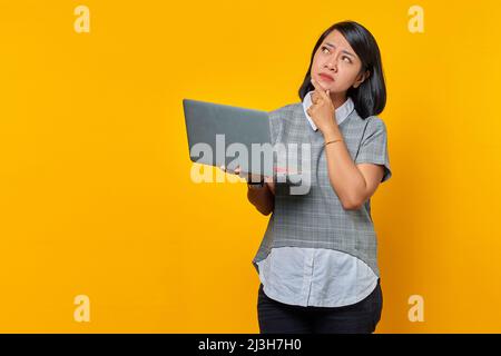 Portrait of beautiful Asian woman holding laptop with serious expression looking up over yellow background Stock Photo