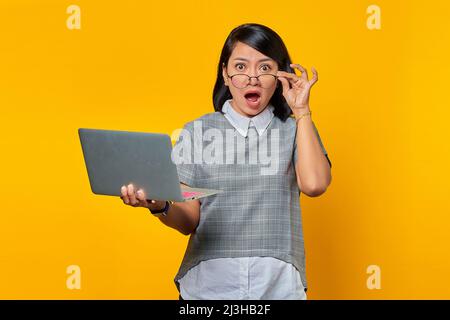 Portrait of surprised young Asian woman holding laptop and touching glasses with finger on yellow background Stock Photo