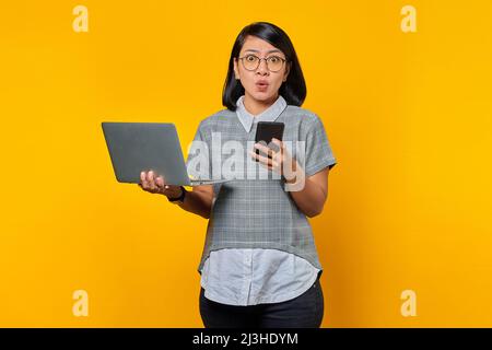 Surprised young woman using laptop and mobile phone while standing isolated on yellow background Stock Photo