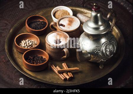 Chai Tea ingredients on a brass plate with tea pot, cinnamon, black pepper, ginger, nutmeg, cardamom, star anise, cloves, black tea, milk and brown sugar Stock Photo