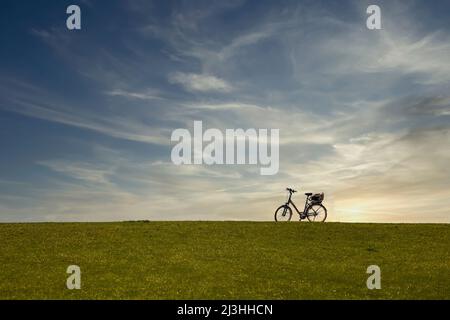 A bicycle stands on top of a dike with evening sky Stock Photo