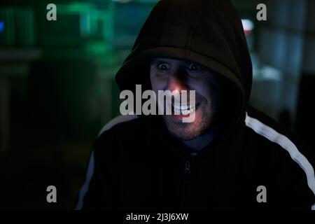 Portrait of mysterious hooded hacker with evil face sitting at desk and breaking into government data servers in dark atmosphere with system codes Stock Photo