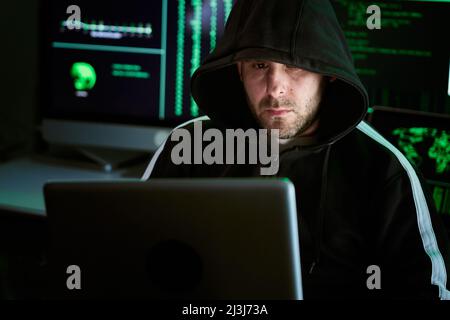 Portrait of criminal hooded hacker sitting at desk and breaking into government or big company data servers in dark atmosphere with system codes Stock Photo
