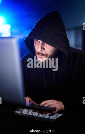 Vertical Portrait of mysterious hooded hacker sitting at desk and breaking into government data servers in dark atmosphere with system codes animation Stock Photo