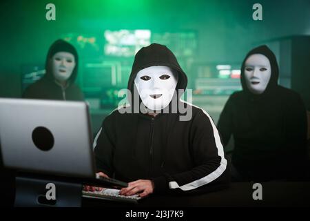 Masked Hacker is Using Computer for Organizing Massive Data Breach Attack on Corporate Servers. They're in Underground Secret Location Surrounded by Stock Photo