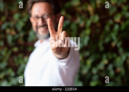 Focus on finger doing victory and peace gesture with cheerful and happy adult man in background. Green nature outdoor leaves defocused. Peaceful peopl Stock Photo