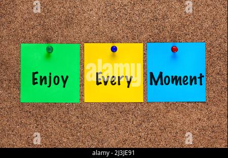 Three colored notes on a corkboard with words Enjoy Every Moment . Close-up Stock Photo