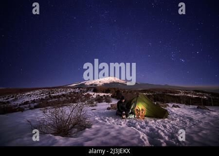 man resting near his illuminated tent on the snow at night under starry sky, on background Etna Mount from Nebrodi Park, Sicily Stock Photo