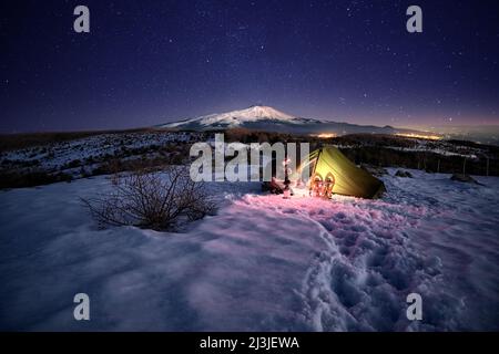 man resting near his illuminated tent on the snow at night under starry sky, on background Etna Mount from Nebrodi Park, Sicily Stock Photo