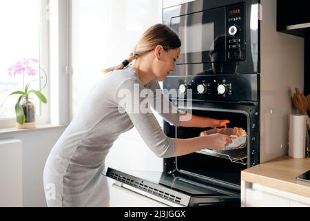 Attractive awesome girl putting baking sheet pie in oven while day light reflects on her body Stock Photo