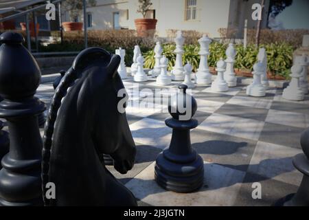 A large outdoor chess game photographed over the shoulder of the black knight looking at the white pawns. Both parties face each other on the battlefi Stock Photo