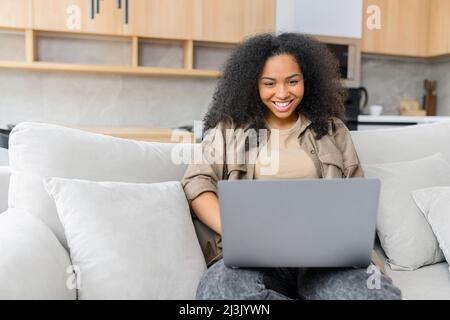 Charming woman spends leisure time in networks on the computer, texting messages, watching movies. Lady works remotely with a laptop sitting on the sofa in cozy living room Stock Photo