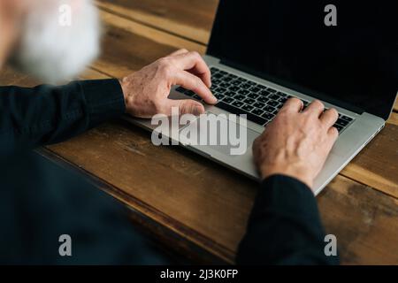 Close-up high-angle view of unrecognizable bearded mature adult male typing on laptop keyboard sitting at table at home office. Stock Photo