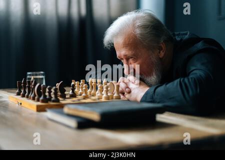 Close-up of pensive gray-haired senior older man thinking game strategy sitting on wooden table with chess board. Stock Photo