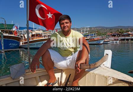 Boatman at Ucagiz, near Kas, Turkey, a young man sitting on the bow of a boat beside a Turkish flag; Ucagiz, Turkey
