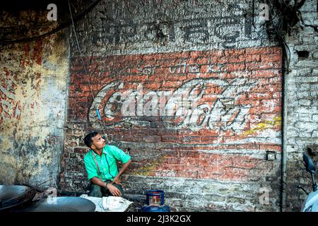 Man sits and looks up at a faded Cola sign painted on a brick wall; Amritsar, Punjab, India Stock Photo