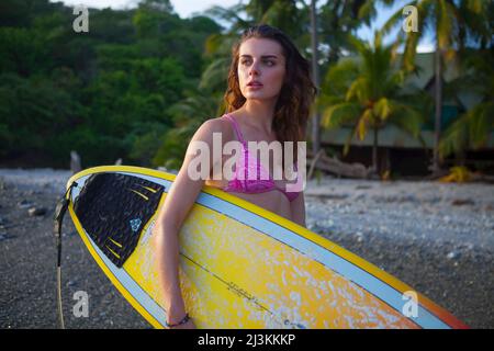A young woman carries a surfboard on a beach. Stock Photo