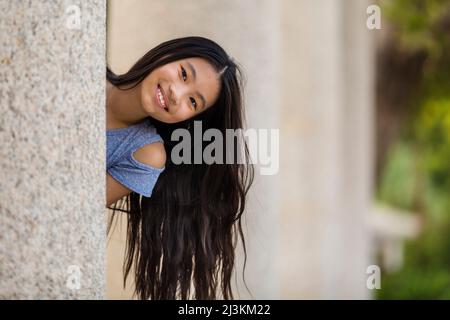 Outdoor portrait of a girl with long, dark hair as she looks out from behind the wall at the camera; Stanley, Hong Kong, China Stock Photo