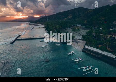 Aerial view of jukung canoes moored at a dock and along the shore of Candidasa Beach with sunlight streaming across the mountaintops under a grey c... Stock Photo