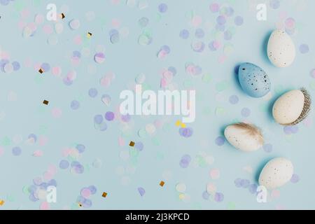 Happy Easter holiday, coming spring concept. Flat lay with white, colorful quail eggs, feather. Blue table background, paper confetti, copy space for ad or text. Easter greeting card, invitation Stock Photo