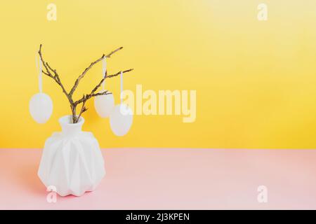 Happy Easter holiday, coming spring concept. Tree branches, flower twigs in white vase or jar with hanging eggs. Yellow and pink table background with copy space for ad or text. Easter home decoration Stock Photo