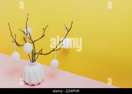 Happy Easter holiday, coming spring concept. Tree branches, flower twigs in white vase or jar with hanging eggs. Yellow and pink table background with copy space for ad or text. Easter home decoration Stock Photo