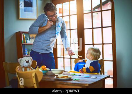 Daddy knows how to multitask. Cropped shot of a single father on a call while feeding his son at home. Stock Photo