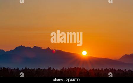 Golden sun setting behind the mountains with sun rays flooding over a forest; Surrey, British Columbia, Canada Stock Photo