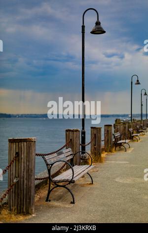 Promenade with benches and lamp posts along a waterfront with rain falling in the distance, Ambleside Beach, West Vancouver Stock Photo