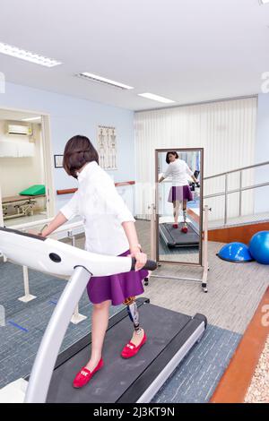 Young woman with leg prosthesis in therapy, walking on a treadmill and looking in the mirror at her reflection as she walks; Bangkok, Thailand Stock Photo