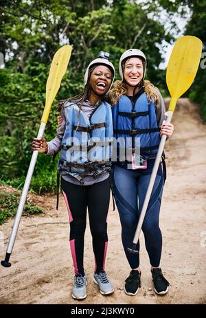 We are ready to go rafting again. Portrait of two cheerful young women wearing protective gear while holding a rowing paddle outside during the day. Stock Photo