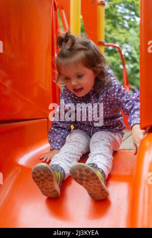 Preschool girl at a playground sitting at the top of a red slide; North Vancouver, British Columbia, Canada Stock Photo