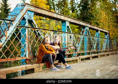 Two sisters sit on a park bridge, one with an acoustic guitar, enjoying music together; Edmonton, Alberta, Canada Stock Photo