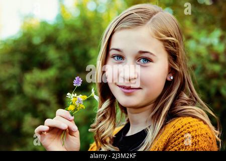 Teenage girl stands outdoors holding a small cluster of colourful wildflowers and looking at the camera; Edmonton, Alberta, Canada
