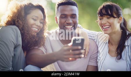 Were starting to trend again guys. Shot of a cheerful group of friends using a cellphone together while relaxing outdoors. Stock Photo