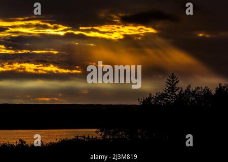 Dramatic colourful sky with sun beams coming down from the sky reflecting on a lake with trees silhouetted in the foreground; Calgary, Alberta, Canada Stock Photo