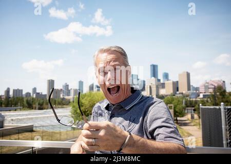 Senior man standing outdoors holds his glasses in his hand to look at the camera with a surprised expression on his face; Edmonton, Alberta, Canada Stock Photo