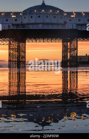 Brighton pier is reflected in the wet sand at low tide as people stroll around on the beach at sunset; Brighton, East Sussex, England Stock Photo