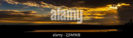 Panorama of a dramatic colourful sky with sun beams coming down from the sky reflecting on a lake with silhouetted hillside; Calgary, Alberta, Canada Stock Photo