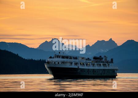 A tour ship cruises Lynn Canal in Southeast Alaska at sunset on its way to Juneau, with Chilkat Mountains beyond; Alaska, United States of America Stock Photo