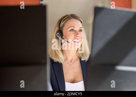 Successful female entrepreneur working on desktop computer while using a headset for conversation in modern office Stock Photo